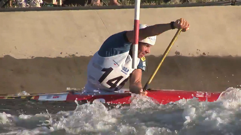 A kayaker wearing a helmet and life vest with the number 14 paddles through rough water in a slalom course, maneuvering around a pole. Splashes of water surround the kayak.