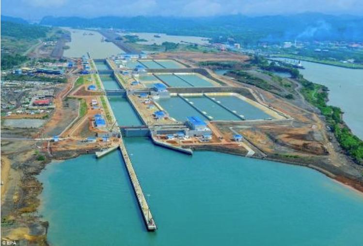 Aerial view of a large canal lock system connecting two bodies of water, showcasing SDVOSB engineering expertise, surrounded by industrial buildings, roads, and greenery, with hills and distant structures in the background.