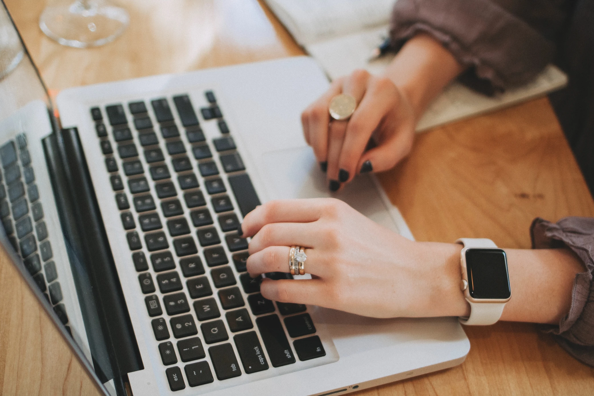 A person wearing a smartwatch and rings types on a laptop keyboard with one hand while using the touchpad with the other. A notebook and pen sit in the background on a wooden desk, reflecting a busy Colorado engineering workspace.