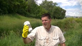 A man wearing a beige shirt and yellow rubber gloves, possibly a civil engineering professional, holds up a white container outdoors, standing in a grassy area with trees and a partly cloudy sky in the background.