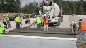 Construction workers in safety vests, overseen by a civil engineering team, pour and spread concrete from a cement truck onto a road surface, using specialized tools and machinery with cars and trees visible in the background.