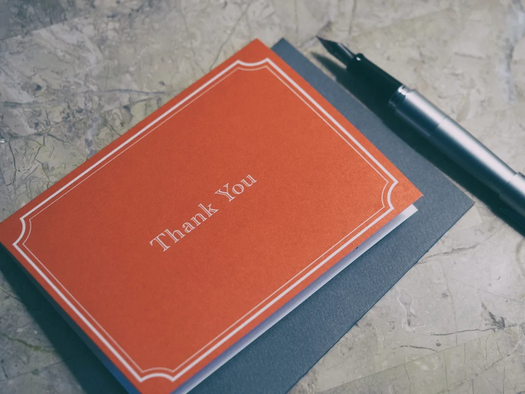 A red thank you card with white trim and lettering sits atop a gray envelope next to a silver fountain pen, on a marble surface—a thoughtful gesture from a Colorado engineering or SDVOSB engineering team.
