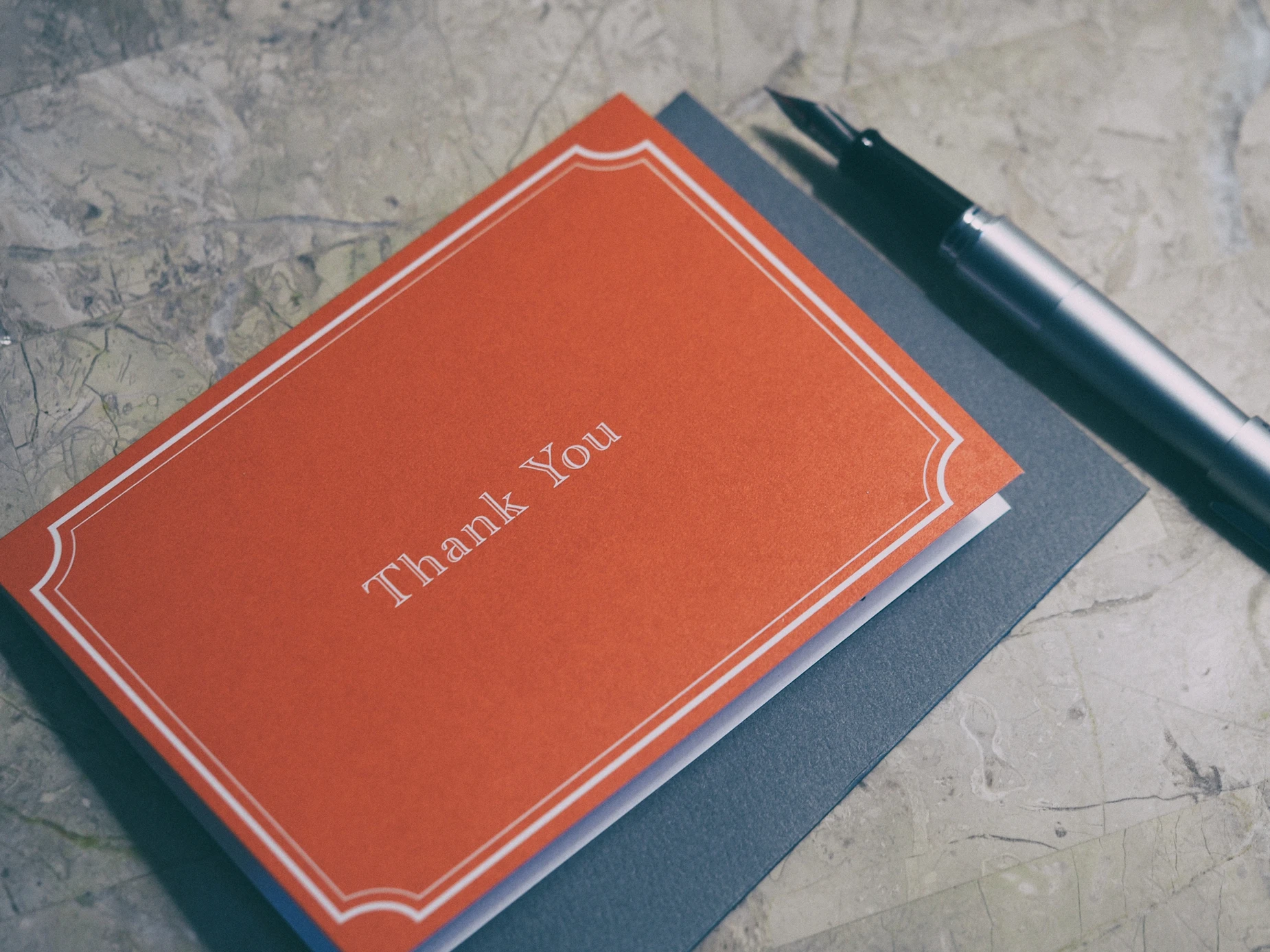 A red thank you card with white trim and lettering sits atop a gray envelope next to a silver fountain pen, on a marble surface—a thoughtful gesture from a Colorado engineering or SDVOSB engineering team.