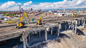 Two large excavators, led by a Denver engineer team, are demolishing a concrete overpass near a rail yard, with city buildings and mountains visible in the background under a partly cloudy sky.
