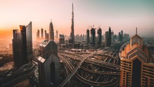 Aerial view of Dubai at sunset, featuring modern skyscrapers shaped by visionary structural engineers, including the Burj Khalifa, and a complex network of highways and interchanges under a warm orange and pink sky.