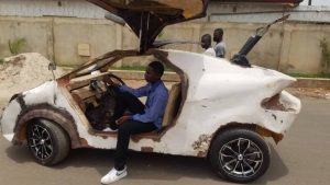A man, possibly a structural engineer, sits in a homemade, rough-looking white car with gullwing doors open, parked on a street. Two people walk by in the background, and a concrete wall with barbed wire lines the street.