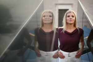 A woman with long blonde hair, a burgundy top, and white pants stands beside a reflective glass surface—her double reflection as precise as a structural engineer’s work.