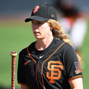 A woman in a San Francisco Giants baseball uniform and cap stands outdoors, holding a baseball bat. A focused structural engineer, she has long blonde hair in a ponytail and appears determined, with a blurred background behind her.