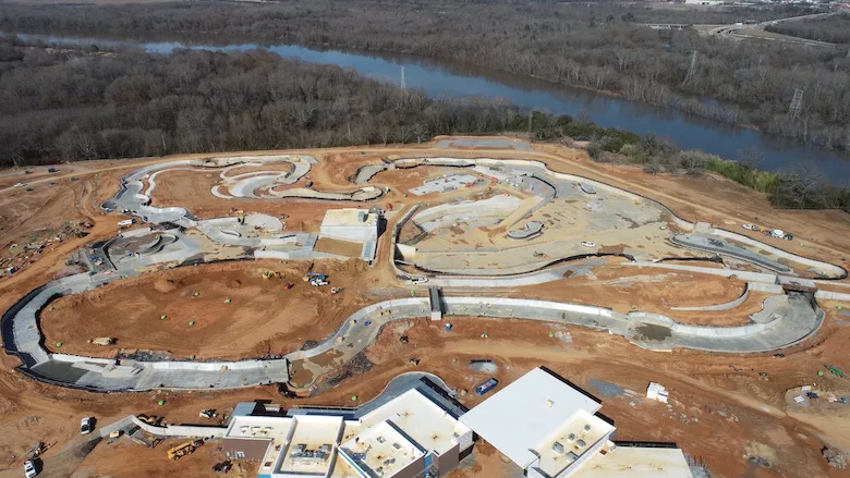 Aerial view of a large construction site with winding concrete water channels and unfinished buildings, surrounded by bare earth. A river and leafless trees are visible in the background on a cloudy day.