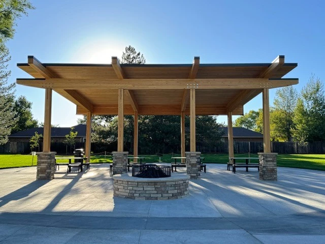 A wooden pavilion with a slatted roof and stone pillars—crafted by a skilled structural engineer—stands in a sunny park, sheltering picnic tables and a central fire pit, all surrounded by green grass and trees.