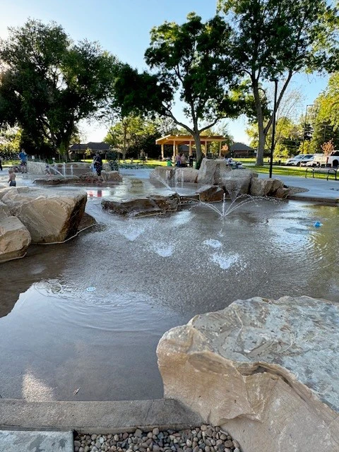 A splash pad with rocks and water fountains, surrounded by trees and grassy park area, designed with a structural engineer’s precision. People play in the background near a yellow pavilion under a clear blue sky.