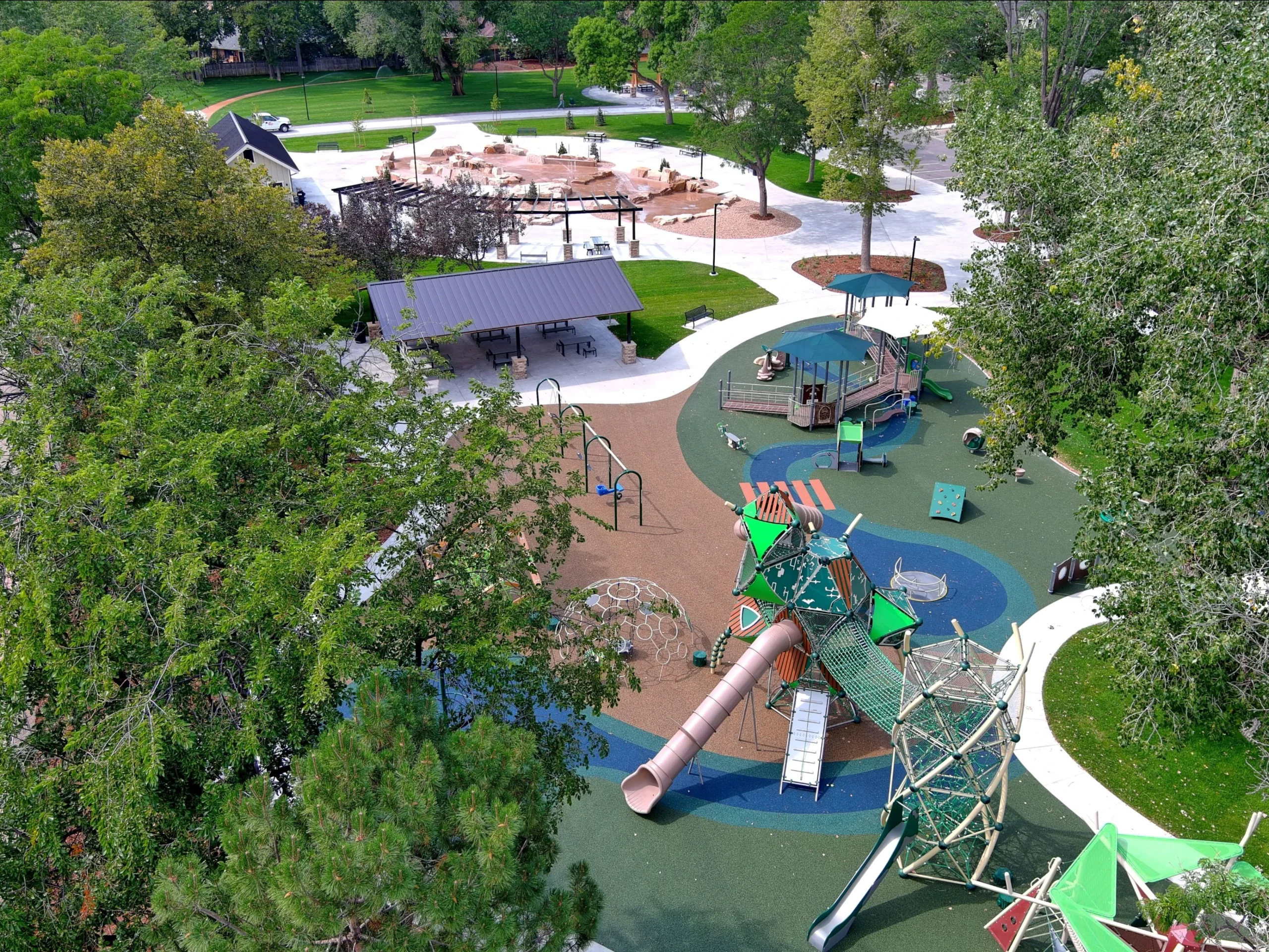 Aerial view of a vibrant outdoor playground with slides, climbing structures, swings, and shaded seating areas—expertly designed with input from a structural engineer—surrounded by large green trees and walking paths.