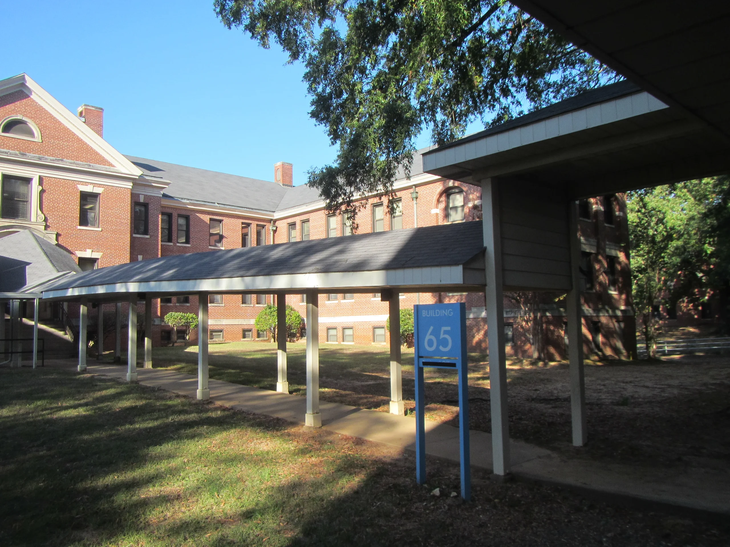 A covered walkway leads to a red-brick building with multiple windows, ideal for a structural engineer. A blue sign by the walkway reads Building 65. Trees and grass surround the area under a clear sky.