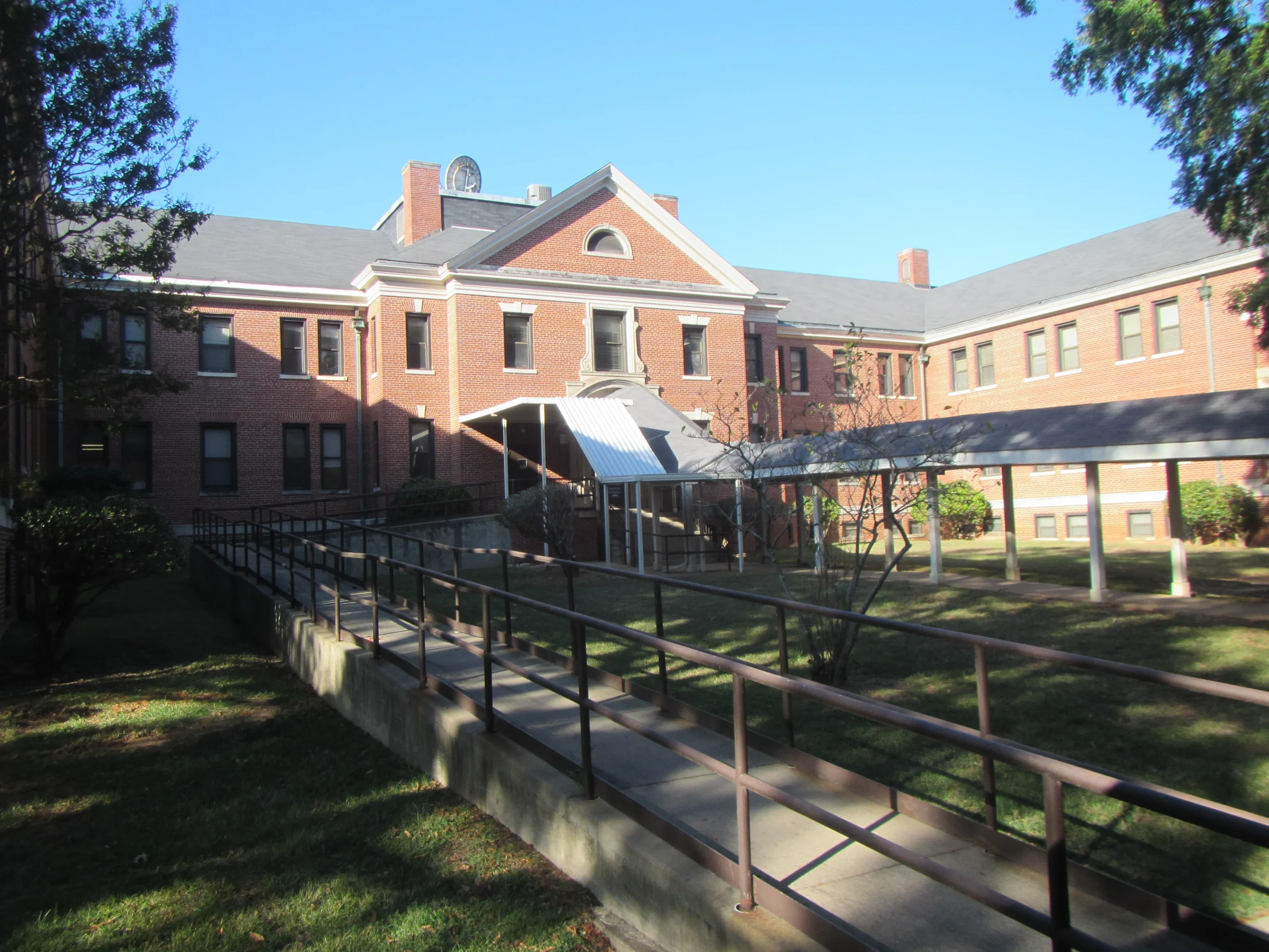 A red-brick building with white trim and a triangular pediment, designed by a skilled Denver engineer, features a covered entrance with a ramp and handrails, surrounded by green lawn and trees on a sunny day.