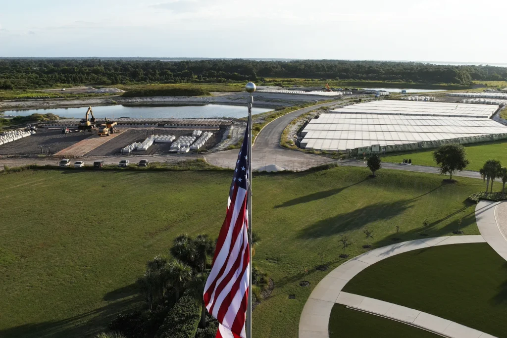 A large American flag waves in the foreground above a green lawn, with a civil engineering construction site, pond, parked vehicles, and stacks of materials visible in the background under a partly cloudy sky.