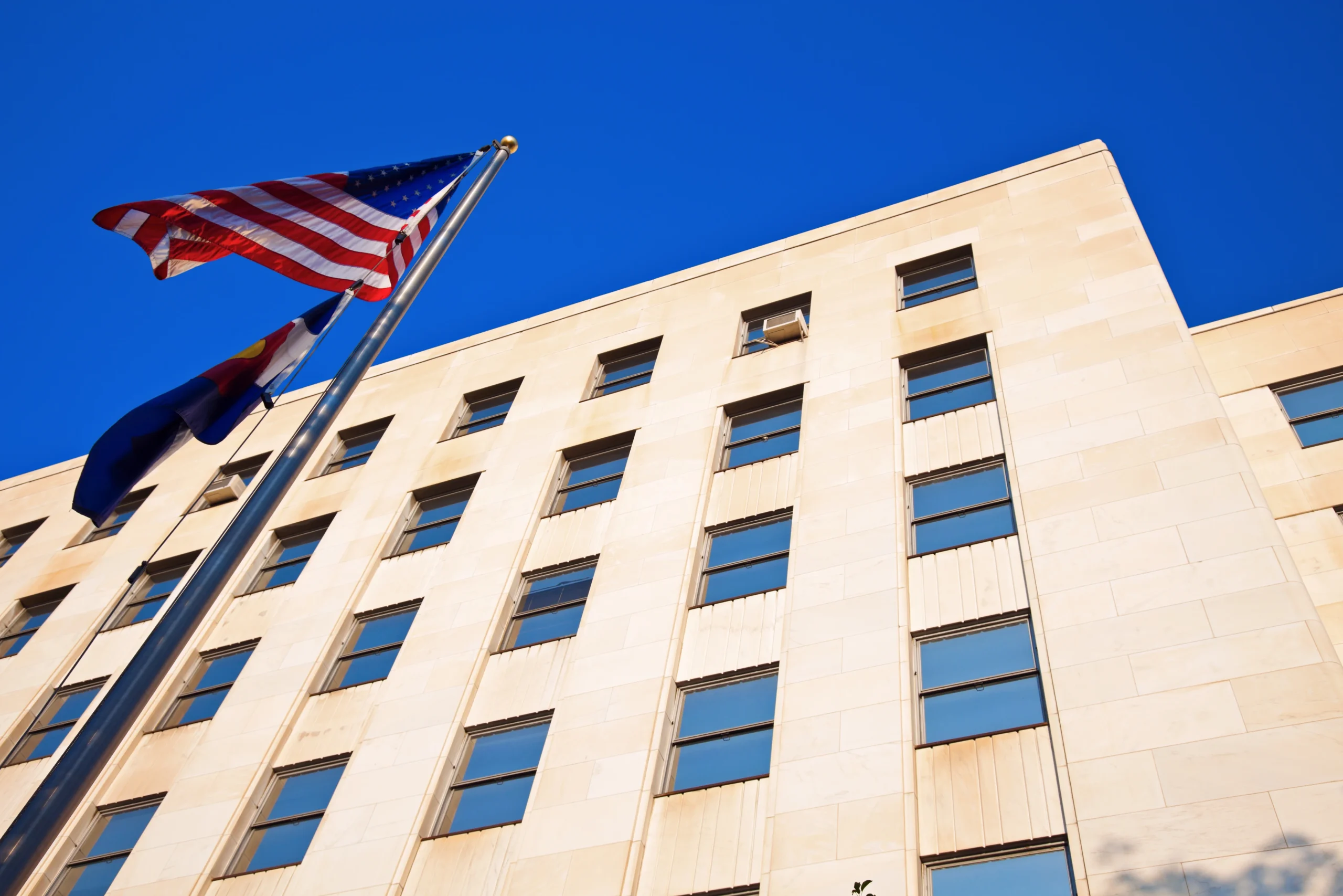 Low-angle view of an American flag in front of a tall beige government or office building, celebrating the work of civil engineering professionals, with many windows and a clear blue sky in the background.