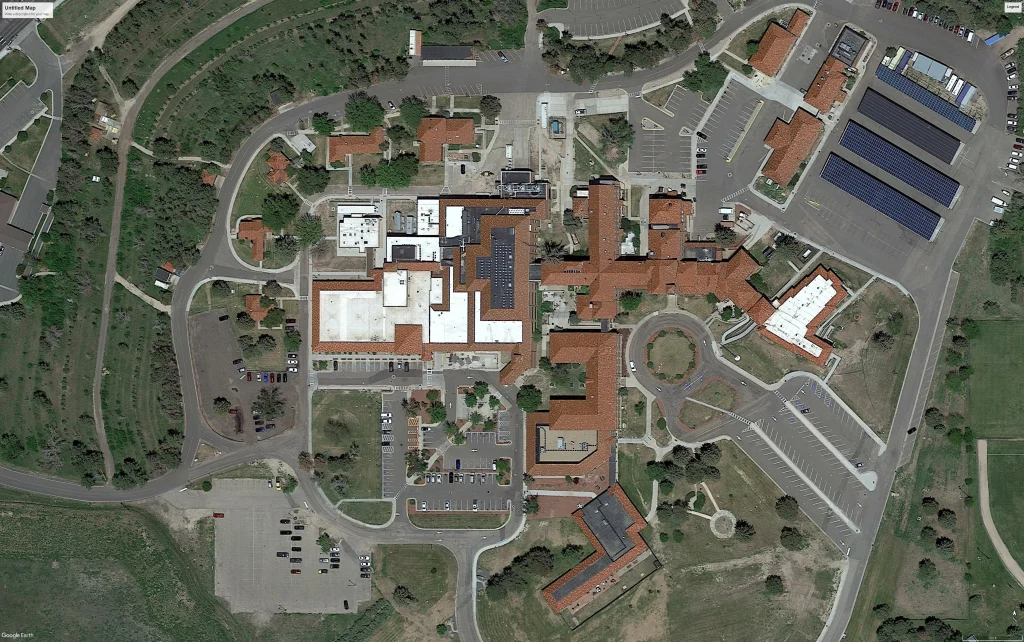 Aerial view of a large building complex with red roofs and white sections, surrounded by parking lots, roads, greenery, and solar panels—reflecting modern civil engineering. Pathways and parked cars are visible throughout the area.