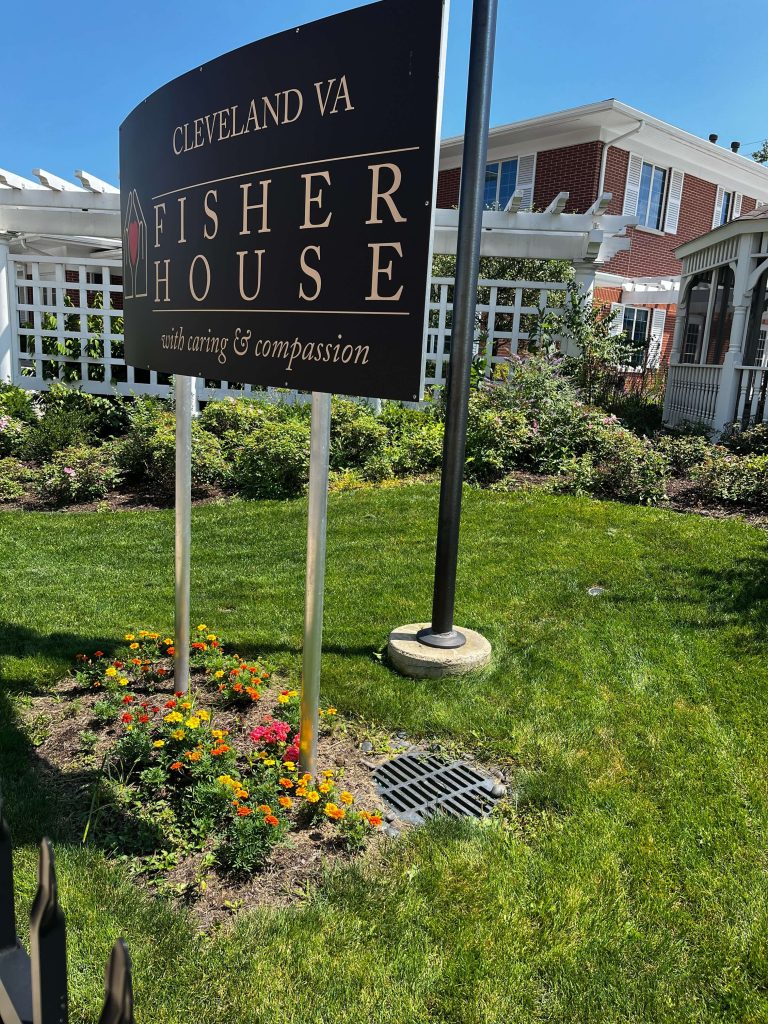 A sign reading Cleveland VA Fisher House with caring & compassion stands on a lawn with yellow and orange flowers, in front of a white fence—built with civil engineering excellence—and a red brick building under a clear blue sky.