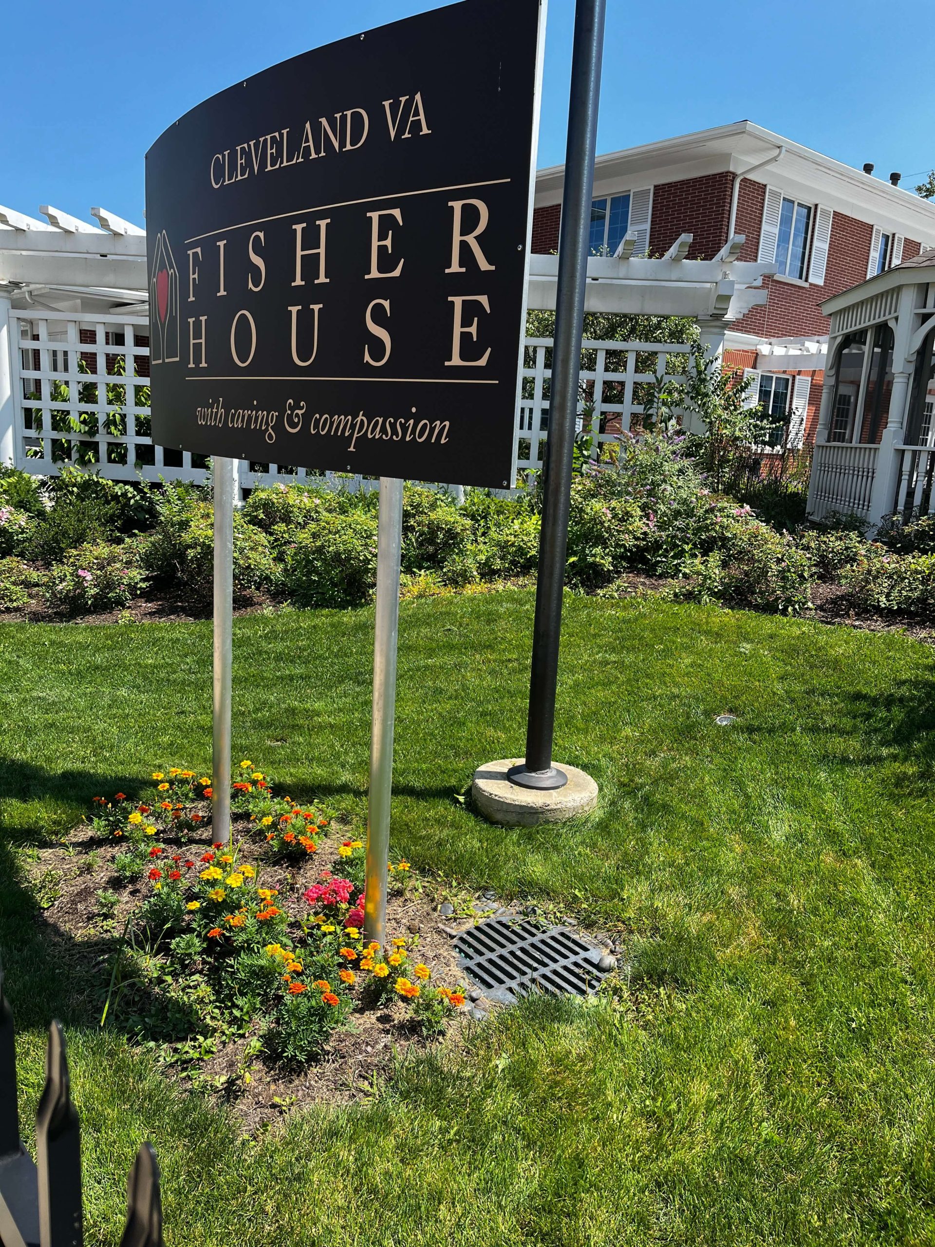 A sign reading Cleveland VA Fisher House with caring & compassion stands on a lawn with yellow and orange flowers, in front of a white fence—built with civil engineering excellence—and a red brick building under a clear blue sky.