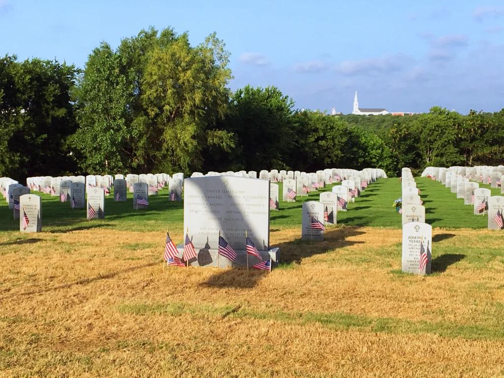 Rows of white headstones in a cemetery, each adorned with small American flags, stand on green and yellow grass. In the distance, trees and a church steeple rise under a blue sky—a scene thoughtfully captured by this Colorado engineering firm.
