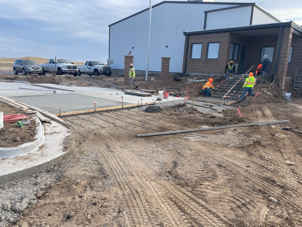 Construction workers in safety gear pour and smooth fresh concrete for a sidewalk and driveway near a building, as SDVOSB engineering professionals oversee the site. Trucks are parked nearby, with dirt and construction materials surrounding the area.