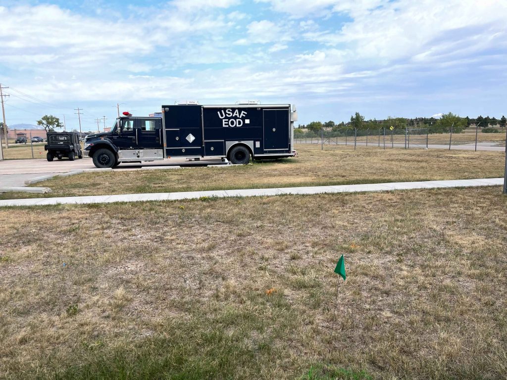 A large black truck labeled USAF EOD is parked on a paved area beside a grassy field, possibly the site of a colorado engineering or civil engineering project, with a small green flag marker in the foreground and a chain-link fence in the background.