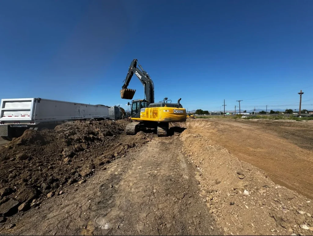A yellow excavator loads dirt into a white dump truck at a construction site under a clear blue sky. Dirt piles and tire tracks mark the ground, while power lines stretch in the background, showcasing real-world civil engineering in action.