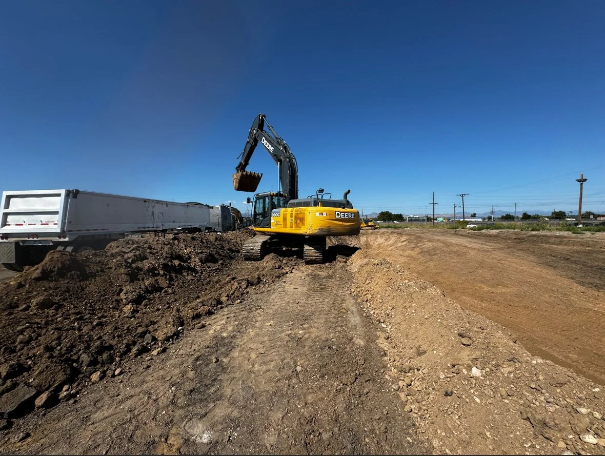 A yellow excavator loads dirt into a white dump truck at a construction site under a clear blue sky. Dirt piles and tire tracks mark the ground, while power lines stretch in the background, showcasing real-world civil engineering in action.