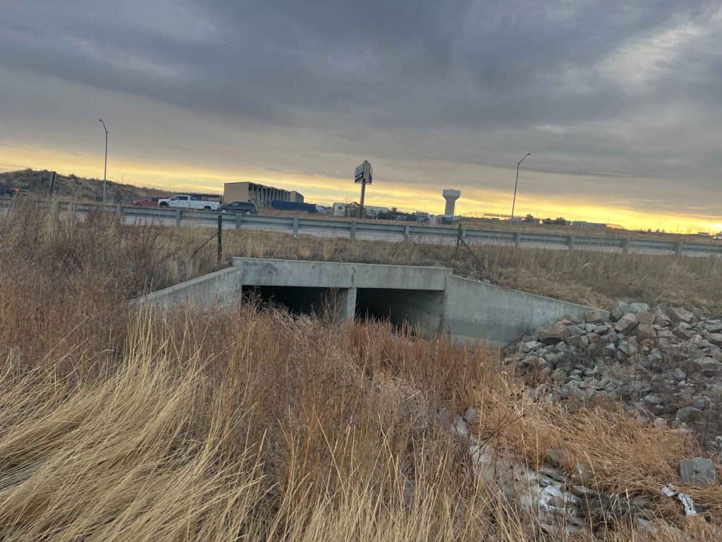 A concrete culvert, designed by a Colorado engineering firm, passes under a highway bordered by dry grass and rocks, with cars on the road and distant buildings beneath a cloudy sunset sky.