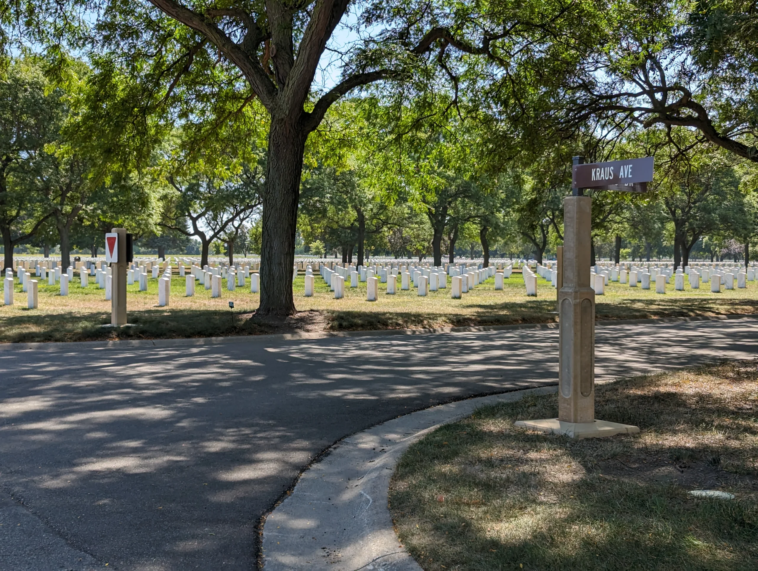Rows of white headstones in a cemetery under large green trees, with a street sign reading “Kraus Ave” on the right and a yield sign on the left near a paved road—evidence of thoughtful civil engineering winding through the peaceful scene.