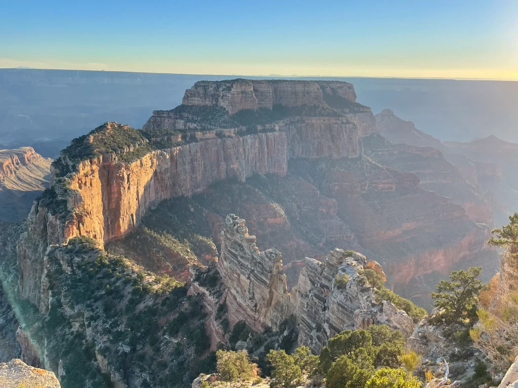 Sunlit cliffs and rock formations of the Grand Canyon rise above deep valleys, with green shrubs in the foreground—an inspiring scene for SDVOSB engineering and civil engineering projects under a hazy blue sky.