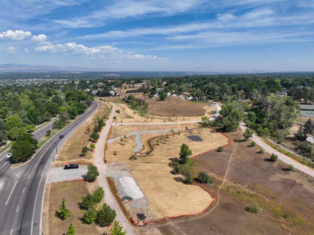 Aerial view of a suburban park under development, with winding paths, a small landscaped area, parked vehicles, and nearby roads surrounded by trees and open fields under a partly cloudy sky.