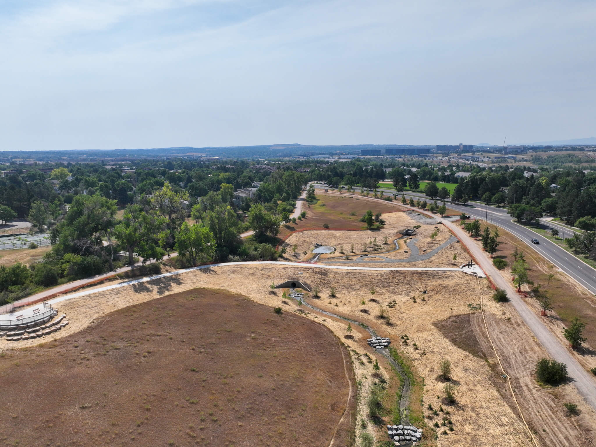 Aerial view of a dry, grassy park with winding paths, sparse trees, and a nearby road under a hazy sky. A circular structure is visible on the left, and a few parked cars are along the paths.