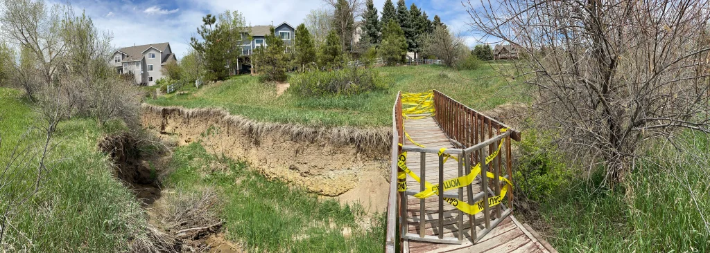 A wooden footbridge with yellow caution tape blocks crosses a grassy creek with eroded banks—an example of civil engineering challenges—while nearby houses and trees rest under a partly cloudy sky.