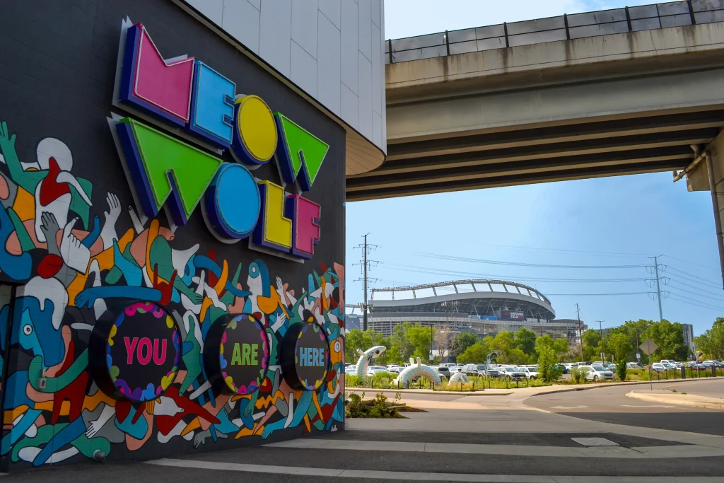 Colorful mural with the words Meow Wolf and You Are Here on a building wall; designed with input from a structural engineer. A stadium and parked cars are visible in the background under an overpass.