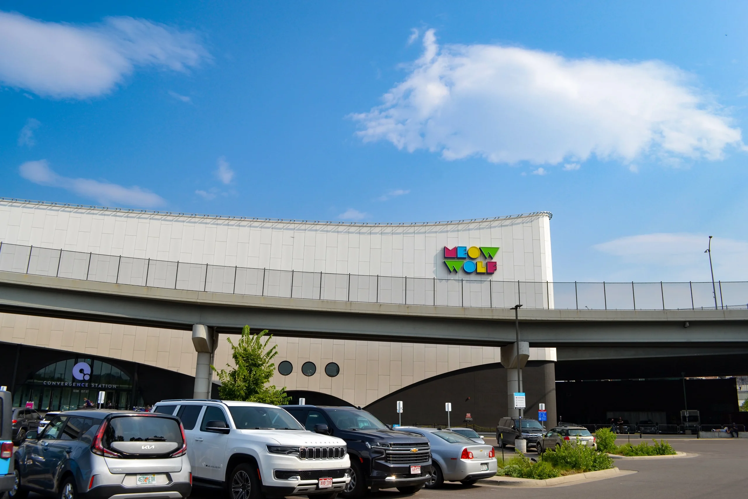 A row of parked cars in front of a modern building with a colorful “Meow Wolf” sign, beneath an elevated roadway designed by a structural engineer, under a bright blue sky with scattered clouds.
