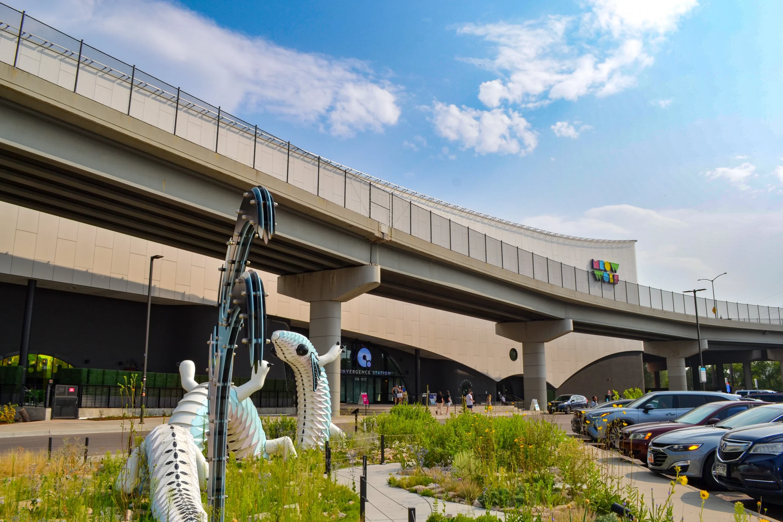 A modern elevated highway, designed by a skilled structural engineer, curves above a building with a colorful sign. In the foreground, a white dinosaur sculpture and wild plants are visible, with cars parked along the right side under partly cloudy skies.