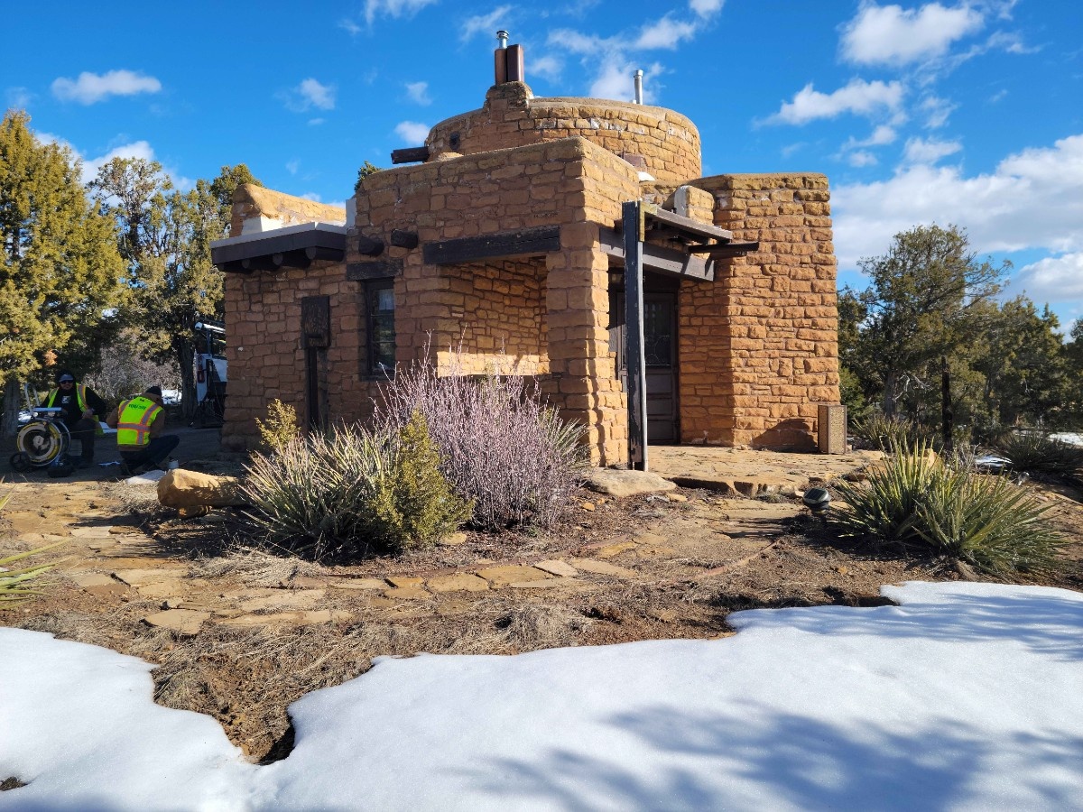 A small, round stone building with a chimney—showcasing Colorado engineering—is surrounded by shrubs and patches of melting snow under a blue sky with scattered clouds. A motorcycle and a person in a yellow vest are partially visible on the left.