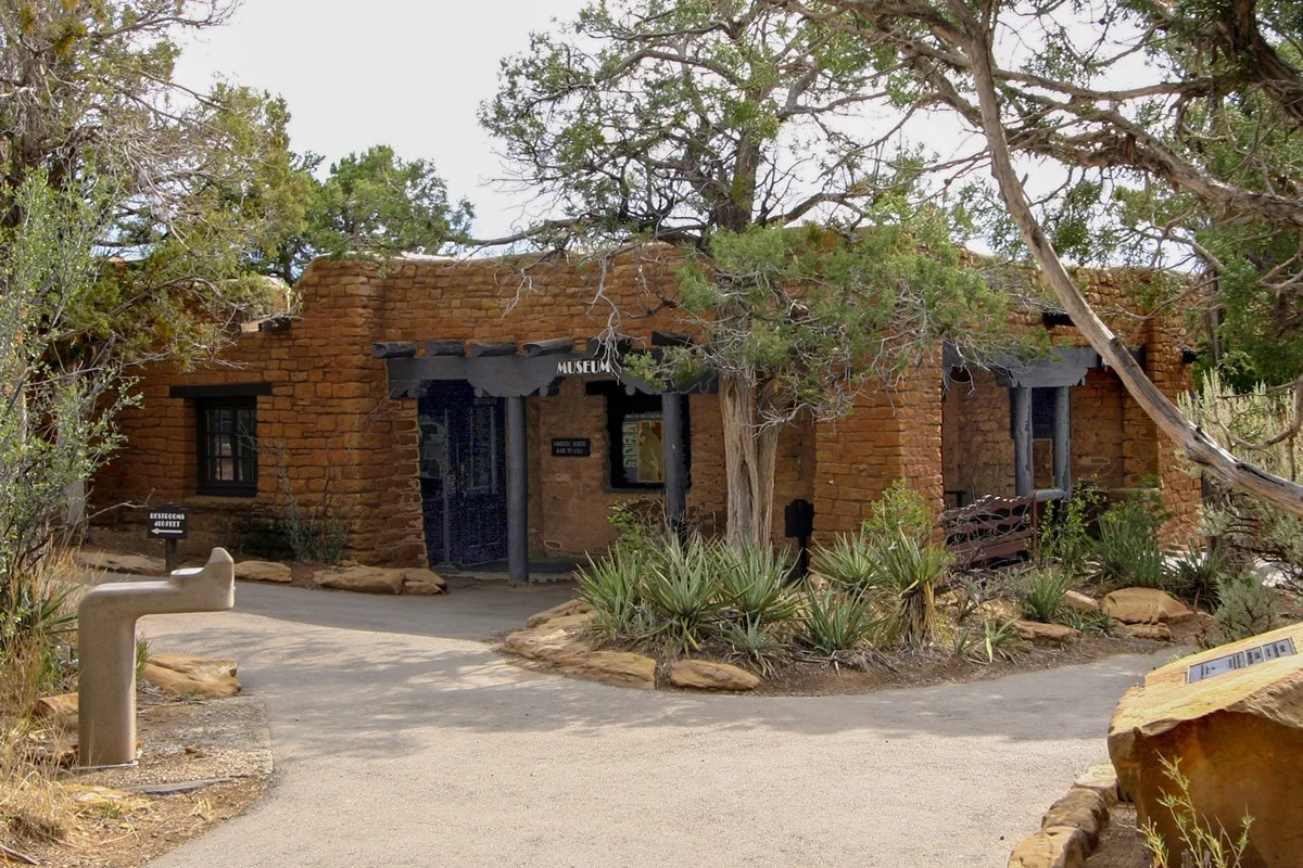 A small adobe building with rustic stone walls and wooden beams sits among desert plants and trees, its sign reading Museum. Designed with expert civil engineering, a paved path leads visitors to the entrance.