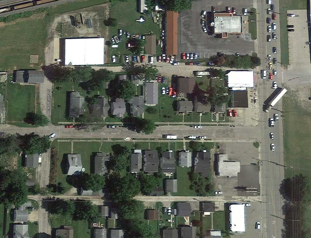 Aerial view of a suburban neighborhood designed with precision, showcasing houses, parked cars, trees, and commercial buildings—an example of civil engineering in action along Denver’s intersecting streets on a sunny day.