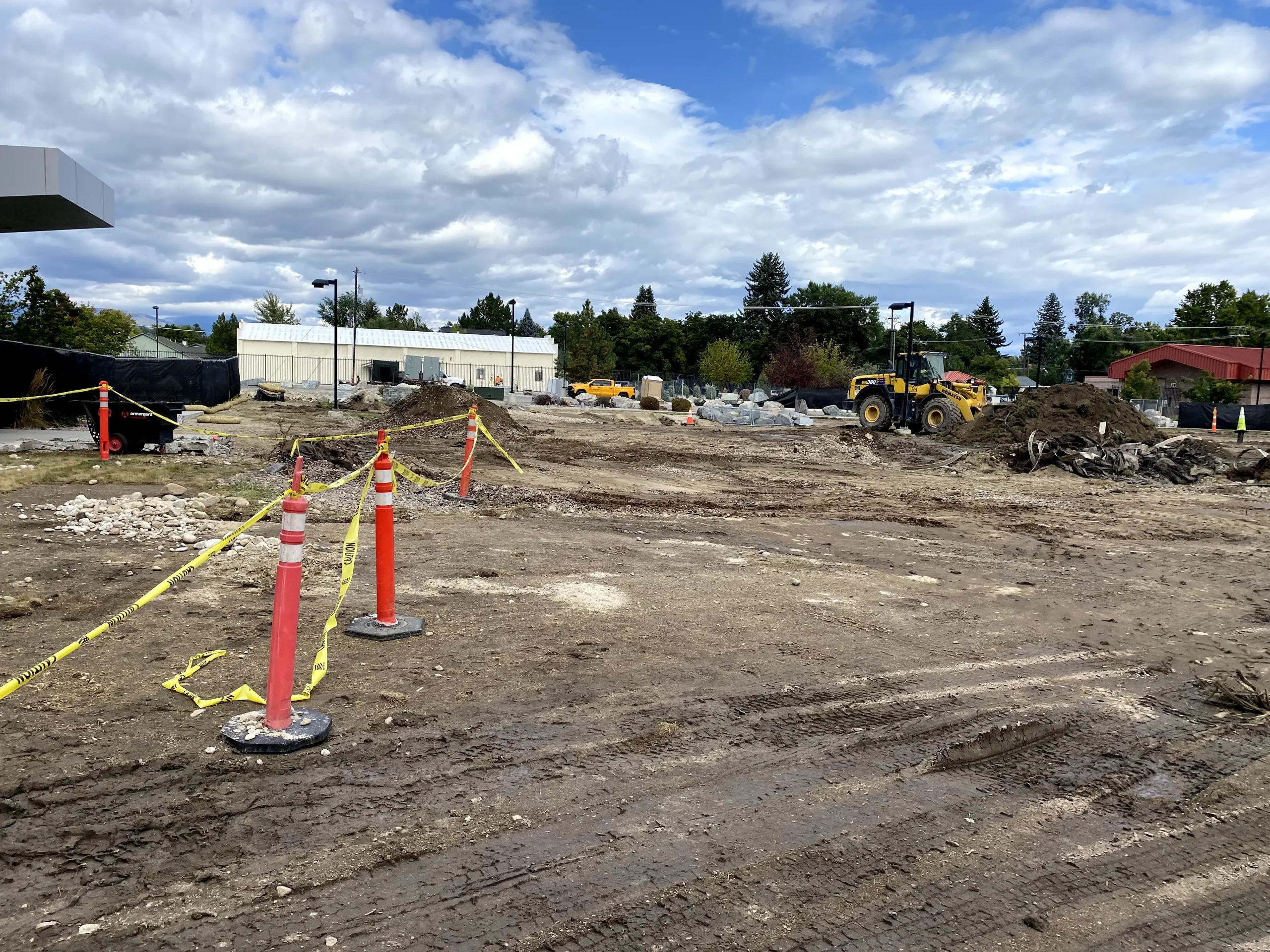 A construction site with dirt ground, scattered debris, orange safety cones, yellow caution tape, and construction vehicles in the background showcases the careful planning of a structural engineer under a partly cloudy sky.