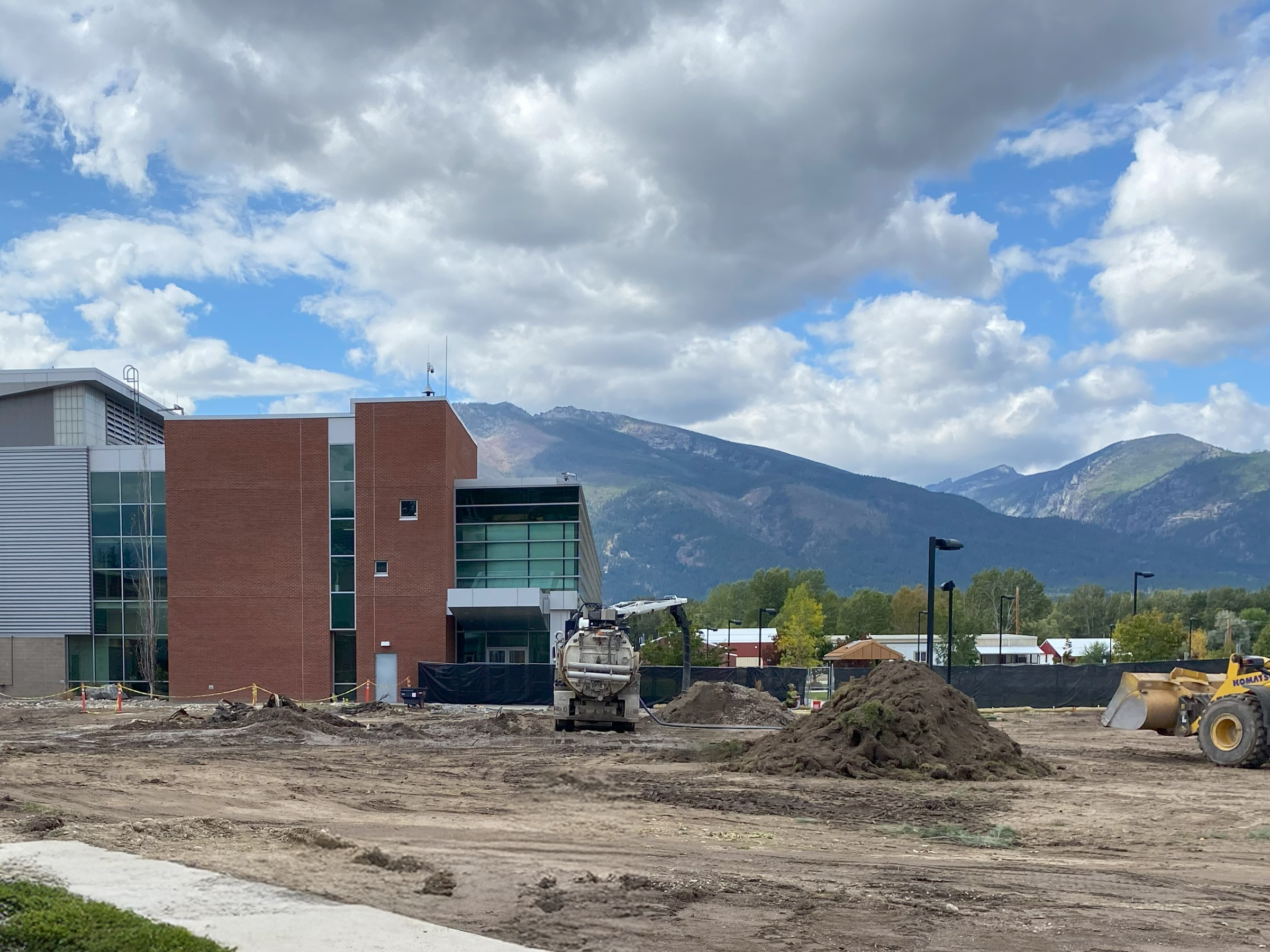 A construction site with piles of dirt and machinery in front of a modern building, set against a backdrop of mountains and a partly cloudy sky.