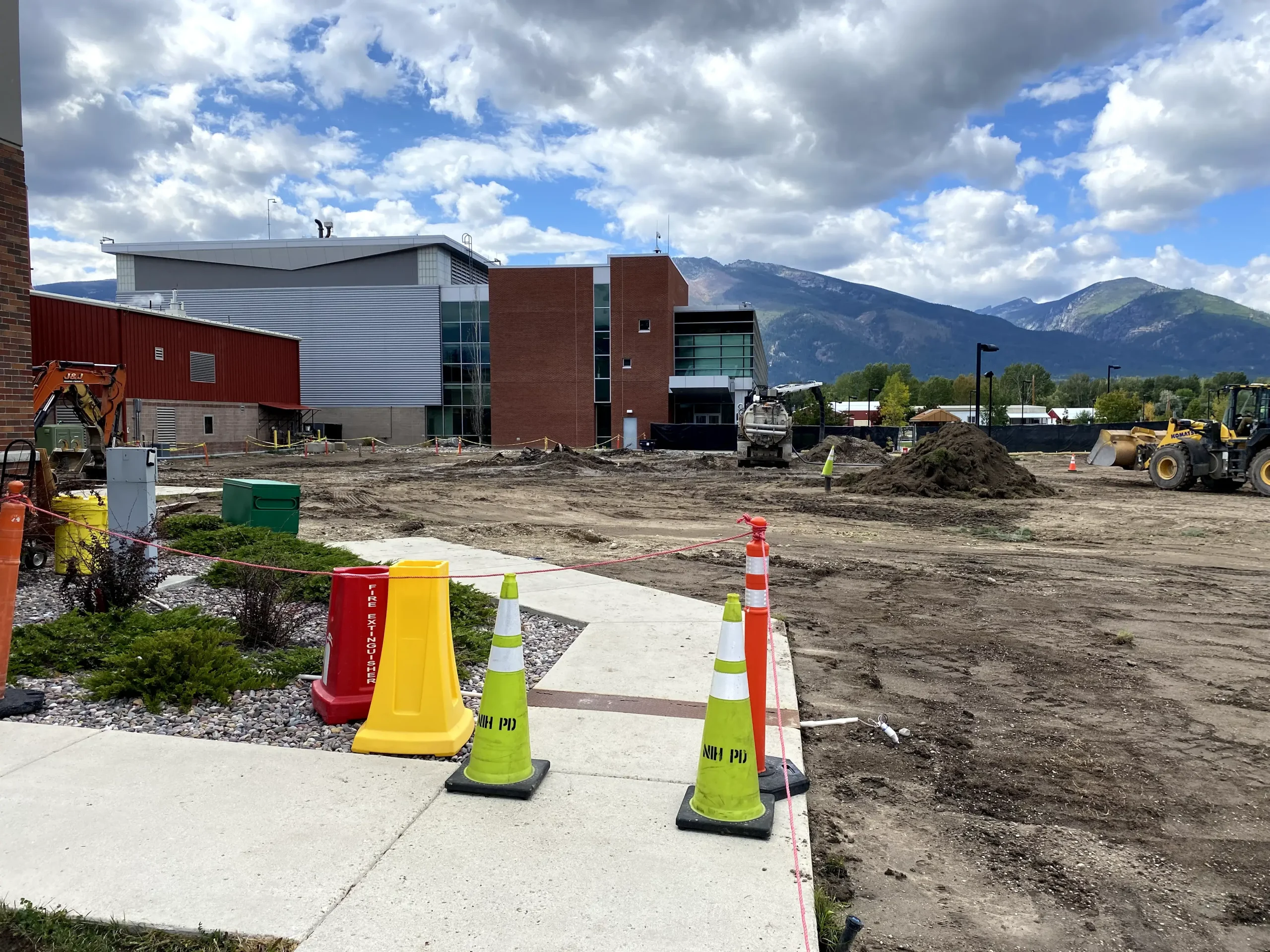 A construction site outside a modern building with mountains in the background. Construction cones and caution signs line a sidewalk, while SDVOSB engineering teams manage dirt piles and machinery under a partly cloudy sky.