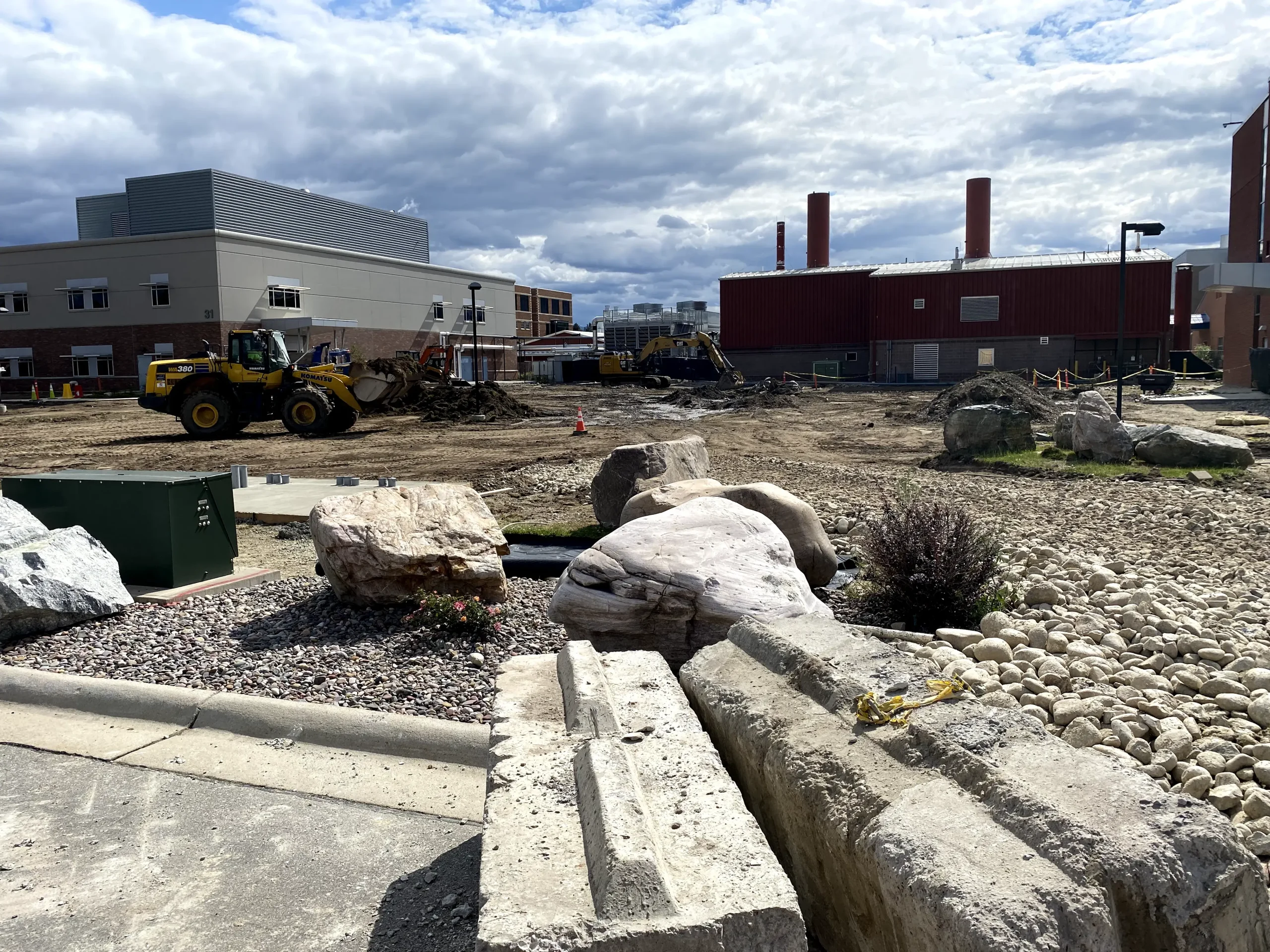 Construction site with heavy machinery, including bulldozers, and dirt piles between two modern buildings. Large rocks and gravel are in the foreground under a partly cloudy sky, showcasing impressive civil engineering at work.