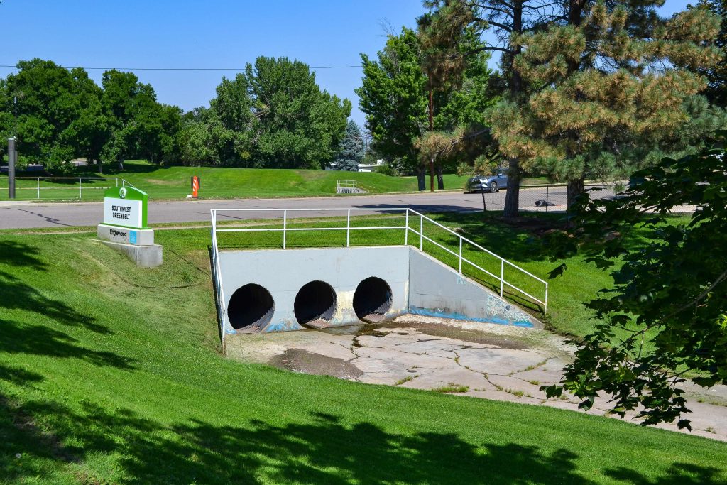 Three large drainage pipes emerge from a concrete structure beneath a sidewalk, surrounded by grassy slopes and trees. A park sign is visible on the left, highlighting colorado engineering expertise in integrating infrastructure with natural landscapes.