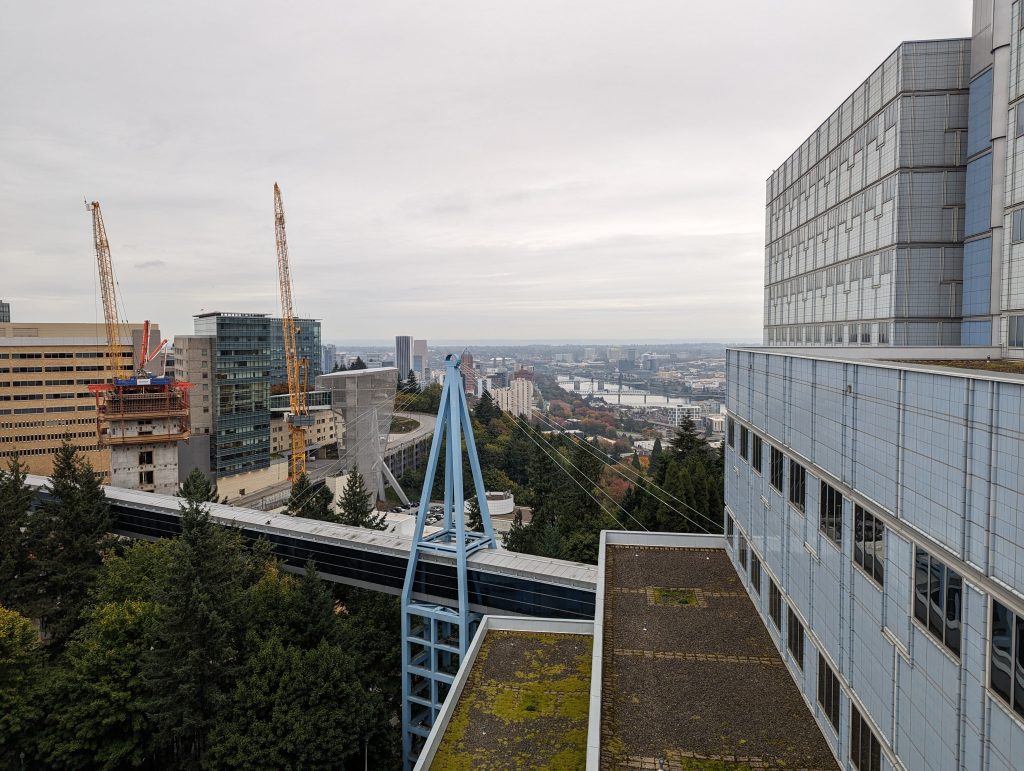 View of a cityscape featuring modern buildings, construction cranes, a cable car system, and a river in the distance—an inspiring scene for any Colorado engineering firm—with cloudy skies and green trees in the foreground.