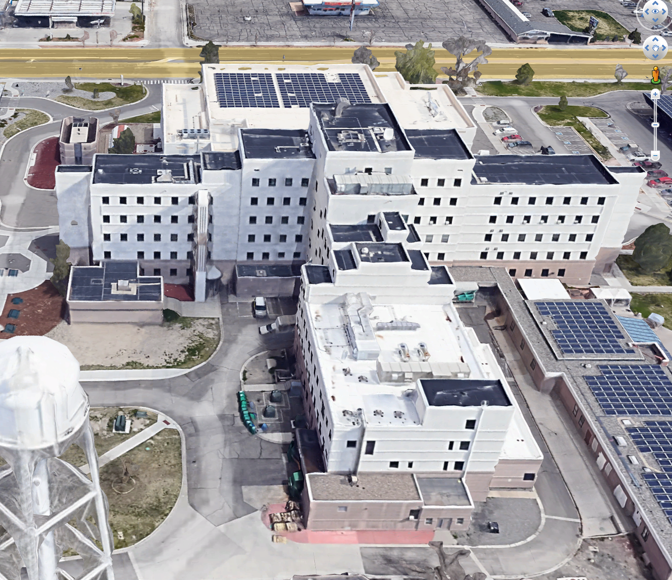 Aerial view of a large, multi-story hospital building with solar panels on the roofs, surrounded by roads, parking areas, and some greenery. A white water tower is visible in the foreground.