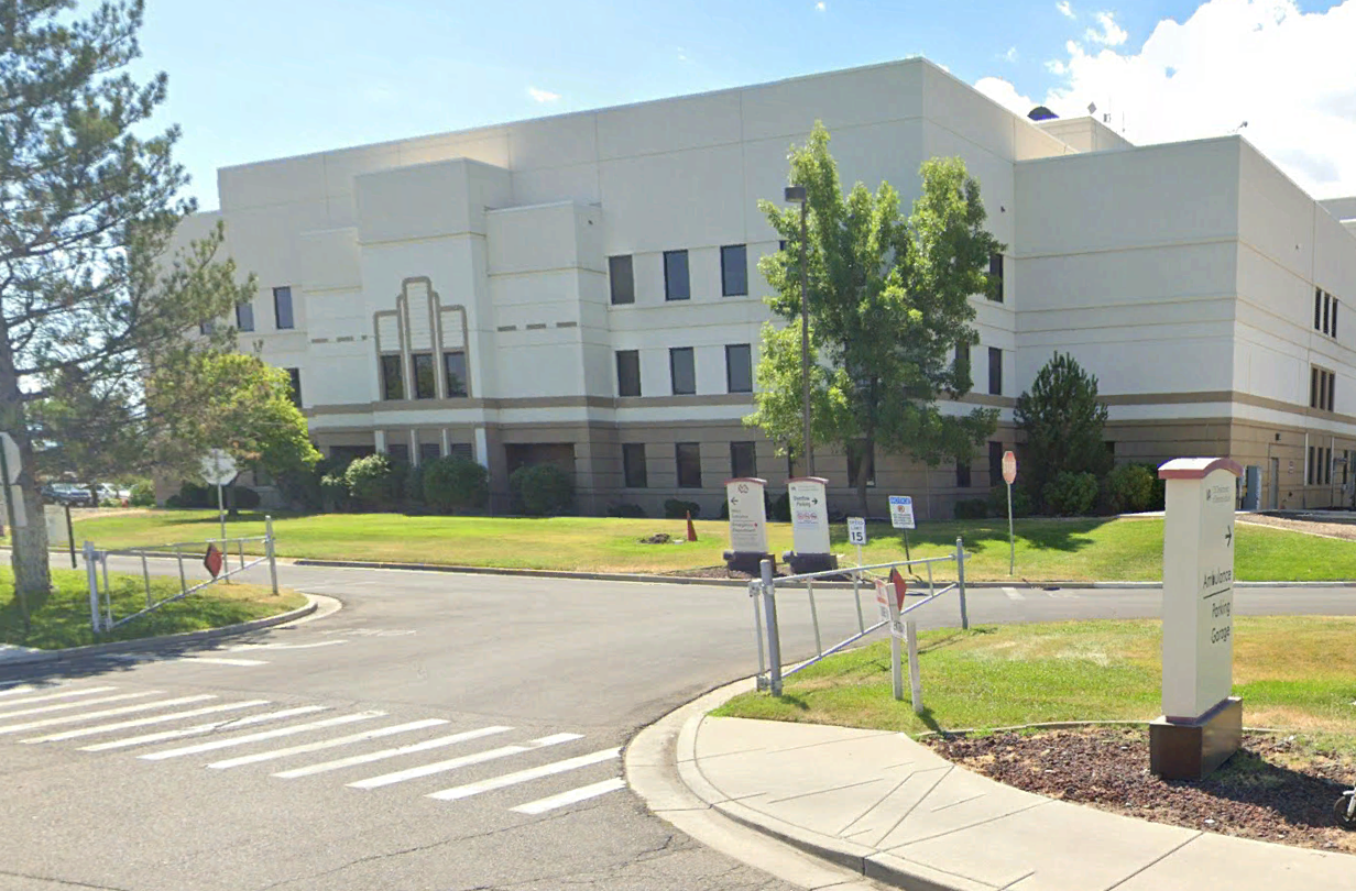 A large, modern, cream-colored building with several windows and a landscaped lawn, surrounded by trees, road signs, and a main entrance driveway under a bright sky.