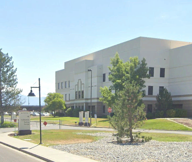 A large white building with multiple windows sits behind trees and a grassy area. A sidewalk, streetlamp, and street signs are visible in the foreground under a clear sky.