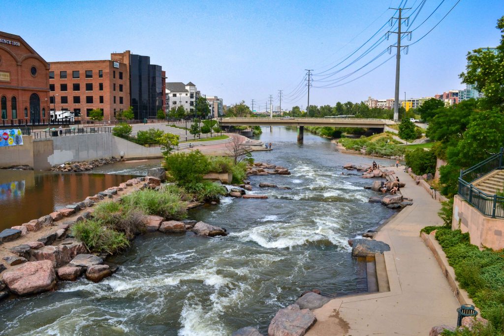 A river with rapids flows through a city, bordered by rocks, greenery, and walkways. People sit near the water; buildings designed with civil engineering expertise, a bridge, and power lines are in the background under a clear sky.